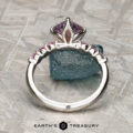 The "Dahlia" ring in platinum with a 2.14-carat Montana sapphire stands upright on a textured surface, with a rough blue-green stone in the background.