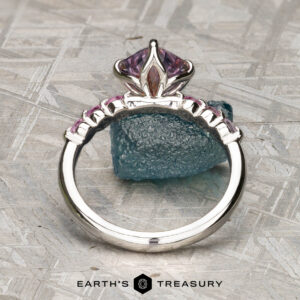 The "Dahlia" ring in platinum with a 2.14-carat Montana sapphire stands upright on a textured surface, with a rough blue-green stone in the background.