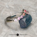 The "Dahlia" band in platinum, adorned with multicolored gemstones, rests on a rough blue stone; a geometric metallic background and the "EARTH'S TREASURY" logo appear below.