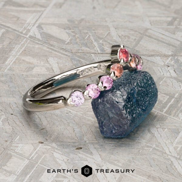 The "Dahlia" band in platinum The "Dahlia" band in platinum, adorned with multicolored gemstones, rests on a rough blue stone; a geometric metallic background and the "EARTH'S TREASURY" logo appear below.