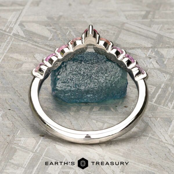 The "Dahlia" band in platinum The "Dahlia" band in platinum, adorned with pink gemstones, stands upright before a rough blue crystal on a textured gray surface. "EARTH'S TREASURY" appears at the bottom.