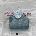 The "Dahlia" ring in 14k white gold with a 1.63-carat Montana sapphire and pink side stones is displayed on rough blue mineral against a textured gray background, with "EARTH'S TREASURY" text at the bottom.