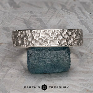 A textured platinum "Luna" band, 6mm wide, with a bubble-like pattern is displayed resting on a rough blue stone, with the "Earth's Treasury" logo below.