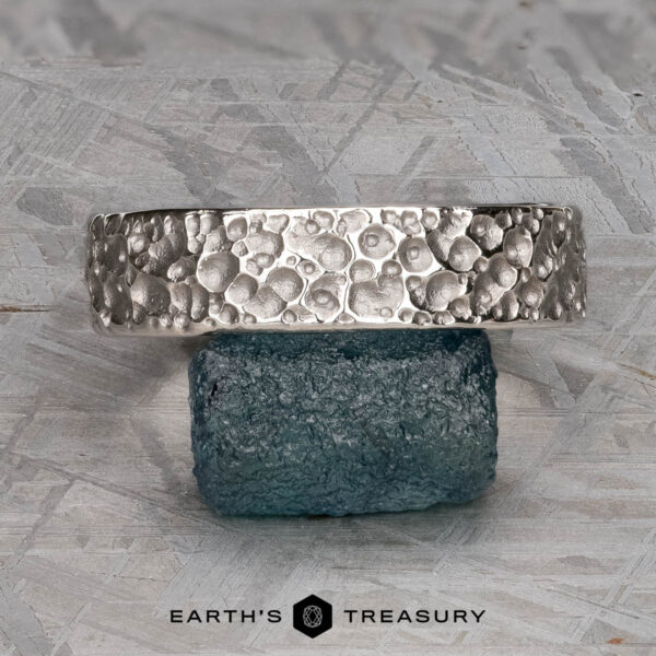 The "Luna" band in platinum, 6mm A textured platinum "Luna" band, 6mm wide, with a bubble-like pattern is displayed resting on a rough blue stone, with the "Earth's Treasury" logo below.