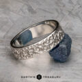 A platinum "Luna" band, 6mm wide with a textured, bubbled surface, is shown leaning against a rough blue gemstone on a gray patterned background. The text "EARTH'S TREASURY" appears below.