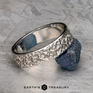 A platinum "Luna" band, 6mm wide with a textured, bubbled surface, is shown leaning against a rough blue gemstone on a gray patterned background. The text "EARTH'S TREASURY" appears below.