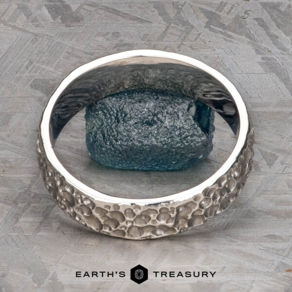 The "Luna" band in platinum, 6mm The "Luna" band in platinum, 6mm, features a textured design encircling a rough blue gemstone, shown on a gray patterned surface with the Earth's Treasury logo below.