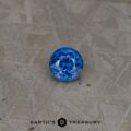 A round, brilliantly cut 2.46-carat Rich Blue Ceylon Sapphire (Heated) is placed on a textured metallic surface. The gemstone reflects light, showcasing its intricate facets and vibrant blue color. The bottom text reads "EARTH'S TREASURY" with a small, intricate logo in between the words.