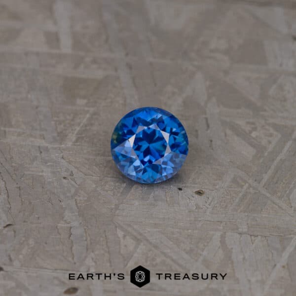 1.76-Carat Medium Blue Ethiopian Sapphire A round, brilliantly cut 2.46-carat Rich Blue Ceylon Sapphire (Heated) is placed on a textured metallic surface. The gemstone reflects light, showcasing its intricate facets and vibrant blue color. The bottom text reads "EARTH'S TREASURY" with a small, intricate logo in between the words.