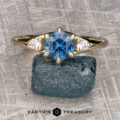 The "Halley" ring in 14k yellow gold features a 1.23-carat hexagonal Montana sapphire flanked by two small white stones, shown on a blue rock with the Earth's Treasury logo—a striking three-stone design.