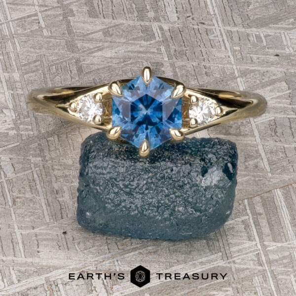 The "Halley" ring in 14k yellow gold with 1.23-Carat Montana Sapphire The "Halley" ring in 14k yellow gold features a 1.23-carat hexagonal Montana sapphire flanked by two small white stones, shown on a blue rock with the Earth's Treasury logo—a striking three-stone design.