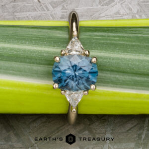 The "Rhiannon" ring in 14k yellow gold with a 2.02-carat Montana Sapphire and two triangular clear stones rests on a green leaf, with "Earth's Treasury" text at the bottom.
