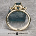 The "Rhiannon" ring in 14k yellow gold, featuring a 2.02-carat Montana Sapphire center and side stones, stands upright before a rough blue crystal on a textured metallic surface. Earth's Treasury logo appears below.