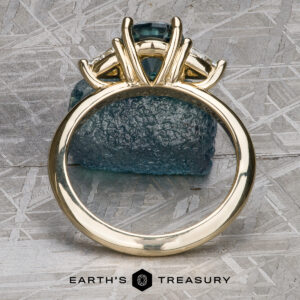 The "Rhiannon" ring in 14k yellow gold, featuring a 2.02-carat Montana Sapphire center and side stones, stands upright before a rough blue crystal on a textured metallic surface. Earth's Treasury logo appears below.