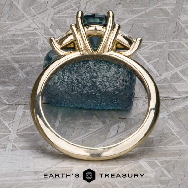 The "Rhiannon" ring in 14k yellow gold with 2.02-Carat Montana Sapphire The "Rhiannon" ring in 14k yellow gold, featuring a 2.02-carat Montana Sapphire center and side stones, stands upright before a rough blue crystal on a textured metallic surface. Earth's Treasury logo appears below.