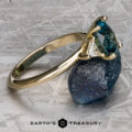 The "Rhiannon" ring in 14k yellow gold with a 2.02-carat Montana sapphire and a white accent stone is showcased on rough blue mineral atop a textured metallic surface.