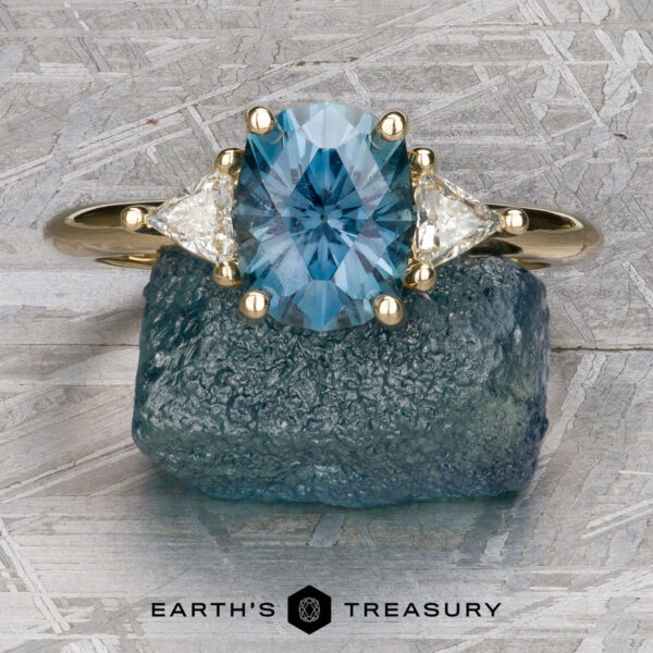The "Rhiannon" ring in 14k yellow gold with 2.02-Carat Montana Sapphire The "Stacked from 51 images. Method=B (R=1,S=1)" ring showcases an oval blue gemstone between two triangular white stones on a gold band, and is beautifully presented atop a rough blue crystal.