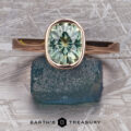 The “Luella” ring in 14k rose gold, featuring a 2.30-carat Montana Sapphire, is displayed atop a rough blue-green stone on a metallic patterned surface with "Earth's Treasury" text below.