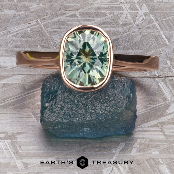 The “Luella” ring in 14k rose gold with 2.30-Carat Montana Sapphire The “Luella” ring in 14k rose gold, featuring a 2.30-carat Montana Sapphire, is displayed atop a rough blue-green stone on a metallic patterned surface with "Earth's Treasury" text below.