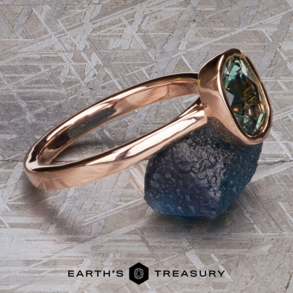 The “Luella” ring in 14k rose gold with 2.30-Carat Montana Sapphire The “Luella” ring in 14k rose gold with a 2.30-carat Montana Sapphire is displayed on a rough blue stone against a textured background, with the "Earth's Treasury" logo at the bottom.