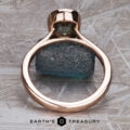 The “Luella” ring in 14k rose gold with a 2.30-carat Montana sapphire stands upright on a rough blue-green stone against a textured gray background, with The Earth's Treasury logo at the bottom.
