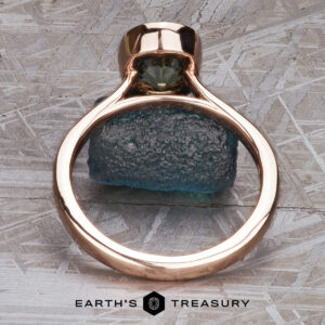 The “Luella” ring in 14k rose gold with a 2.30-carat Montana sapphire stands upright on a rough blue-green stone against a textured gray background, with The Earth's Treasury logo at the bottom.