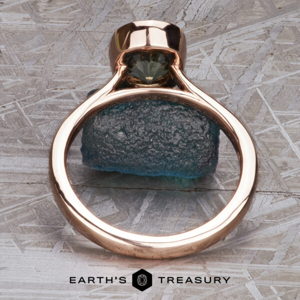 The “Luella” ring in 14k rose gold with 2.30-Carat Montana Sapphire The “Luella” ring in 14k rose gold with a 2.30-carat Montana sapphire stands upright on a rough blue-green stone against a textured gray background, with The Earth's Treasury logo at the bottom.