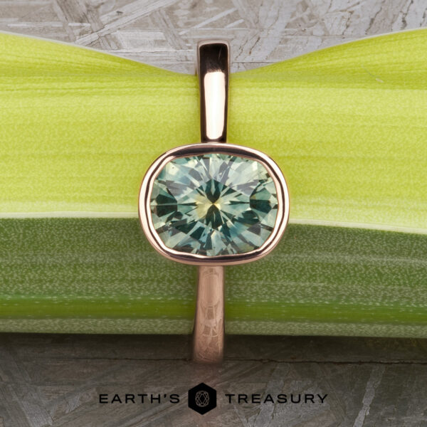 The “Luella” ring in 14k rose gold with 2.30-Carat Montana Sapphire The “Luella” engagement ring in 14k rose gold with a 2.30-carat Montana Sapphire is shown upright against a green-striped background, featuring the "Earth's Treasury" logo and text below.