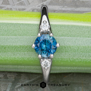 The "Halley" ring in 18k white gold showcases a 0.91-carat Montana sapphire at its center, flanked by two smaller clear stones in a classic three-stone design, displayed on a green and white background.