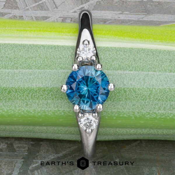 The "Halley" ring in 18k white gold with 0.91-carat Montana sapphire The "Halley" ring in 18k white gold showcases a 0.91-carat Montana sapphire at its center, flanked by two smaller clear stones in a classic three-stone design, displayed on a green and white background.
