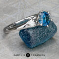 The "Halley" ring in 18k white gold featuring a 0.91-carat Montana sapphire rests on a blue rough stone atop a patterned surface. The "EARTH'S TREASURY" logo is shown at the bottom.