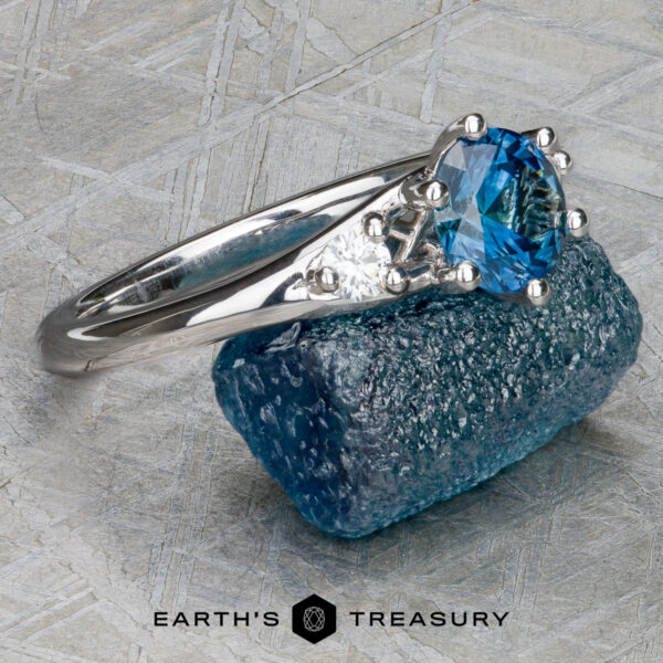 The "Halley" ring in 18k white gold with 0.91-carat Montana sapphire The "Halley" ring in 18k white gold featuring a 0.91-carat Montana sapphire rests on a blue rough stone atop a patterned surface. The "EARTH'S TREASURY" logo is shown at the bottom.