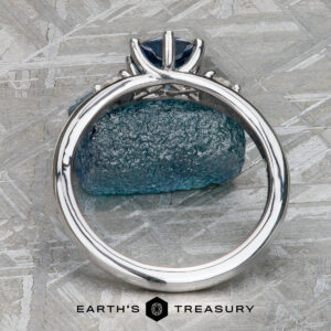 The "Halley" ring in 18k white gold with a 0.91-carat Montana sapphire stands upright before a rough blue mineral, branded "Earth's Treasury," its celestial-inspired design radiating elegance.