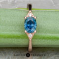 The "Niamh" ring in 14k rose gold features a 0.95-carat oval Montana sapphire center and two small white accent stones, shown on a green surface with the Earth's Treasury logo at the bottom.