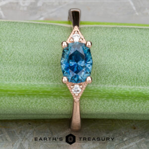 The "Niamh" ring in 14k rose gold features a 0.95-carat oval Montana sapphire center and two small white accent stones, shown on a green surface with the Earth's Treasury logo at the bottom.