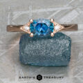 The "Niamh" ring in 14k rose gold, featuring a 0.95-carat Montana Sapphire and smaller clear stones, is displayed on a rough blue stone surface with the "Earth's Treasury" text and logo below.
