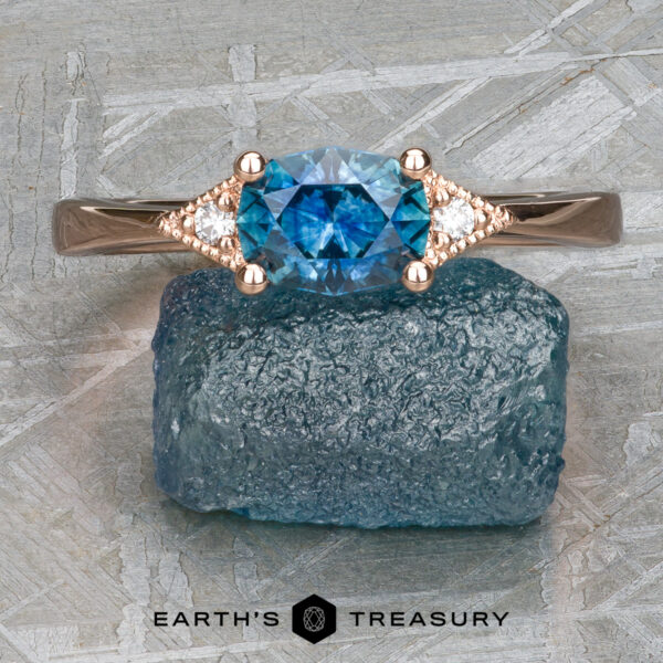 The "Niamh" ring in 14k rose gold with 0.95-Carat Montana Sapphire The "Niamh" ring in 14k rose gold, featuring a 0.95-carat Montana Sapphire and smaller clear stones, is displayed on a rough blue stone surface with the "Earth's Treasury" text and logo below.