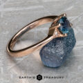 A "Niamh" ring in 14k rose gold with a 0.95-carat Montana sapphire is shown on a rough blue stone surface, featuring the Earth's Treasury logo at the bottom.