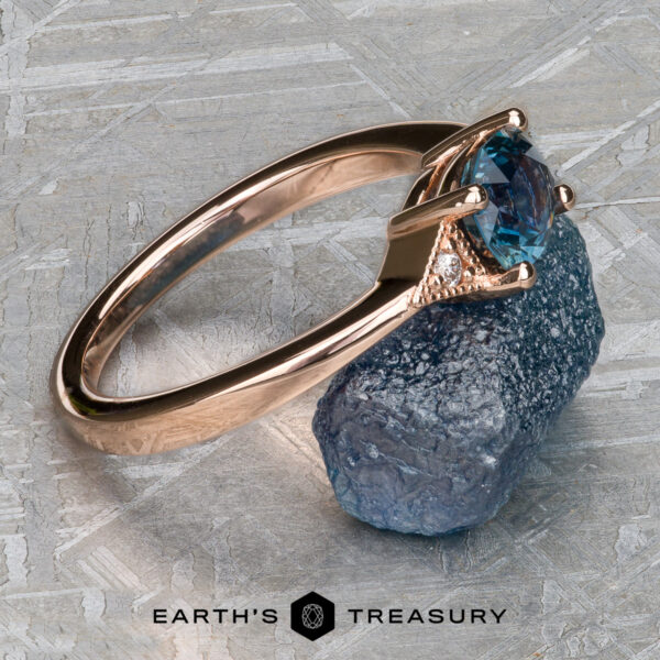 The "Niamh" ring in 14k rose gold with 0.95-Carat Montana Sapphire A "Niamh" ring in 14k rose gold with a 0.95-carat Montana sapphire is shown on a rough blue stone surface, featuring the Earth's Treasury logo at the bottom.