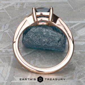 A "Niamh" ring in 14k rose gold with a 0.95-carat Montana sapphire is displayed upright on a rough blue mineral specimen, with the "Earth's Treasury" logo and text below.