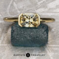 The “Larisa” ring in 18k yellow gold with a 1.35-carat Montana sapphire is showcased on a rough blue-green stone, with "Earth's Treasury" branding below, highlighting its timeless simplicity and natural beauty.