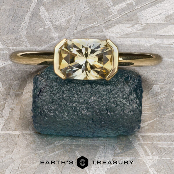 The “Larisa” ring in 18k yellow gold with 1.35-Carat Montana sapphire The “Larisa” ring in 18k yellow gold with a 1.35-carat Montana sapphire is showcased on a rough blue-green stone, with "Earth's Treasury" branding below, highlighting its timeless simplicity and natural beauty.