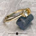 The “Larisa” ring in 18k yellow gold with a 1.35-carat Montana sapphire rests on a rough blue stone atop a textured gray surface, with the "Earth's Treasury" logo at the bottom.