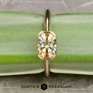 The “Larisa” ring in 18k yellow gold with a 1.35-carat Montana sapphire is displayed upright against a green textured background with the "Earth's Treasury" logo at the bottom, highlighting its elegance.