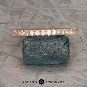 The petite pave diamond band in 14k rose gold, hammered and brushed, rests on a rough dark stone with a textured gray background. "Earth's Treasury" logo appears below.