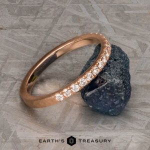 The petite pave diamond band in 14k rose gold, hammered and brushed, is displayed leaning against a rough dark stone on a textured surface, with the "EARTH'S TREASURY" logo beneath.