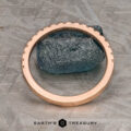 A hammered and brushed 14k rose gold petite pavé diamond band stands upright around a rough blue stone on a patterned metal surface. Text at the bottom reads "Earth's Treasury.