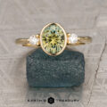 The "Evadne" ring in 18K yellow gold showcases a 1.40-carat oval Montana sapphire between two diamonds, displayed upright on a rough dark green stone.