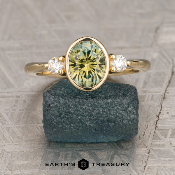 The "Evadne" ring in 18K yellow gold with 1.40-carat Montana sapphire The "Evadne" ring in 18K yellow gold showcases a 1.40-carat oval Montana sapphire between two diamonds, displayed upright on a rough dark green stone.