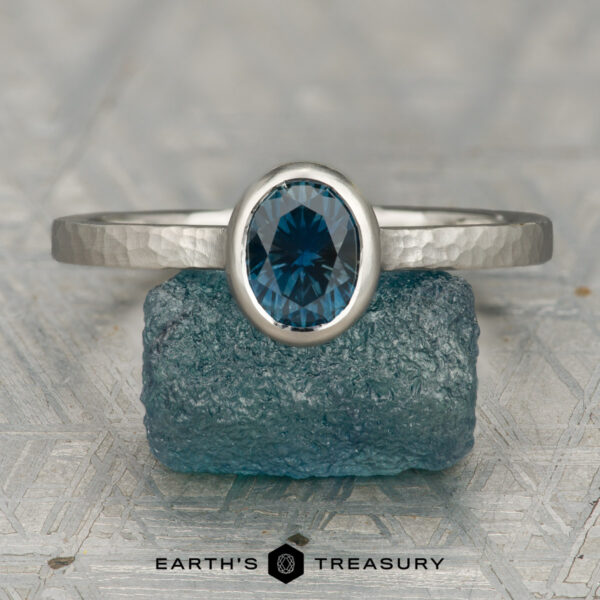 The "Luella" ring in platinum with 1.15-carat Montana sapphire The "Luella" ring in platinum with a 1.15-carat Montana sapphire is displayed upright on a rough blue stone, with the "Earth's Treasury" logo and text below.